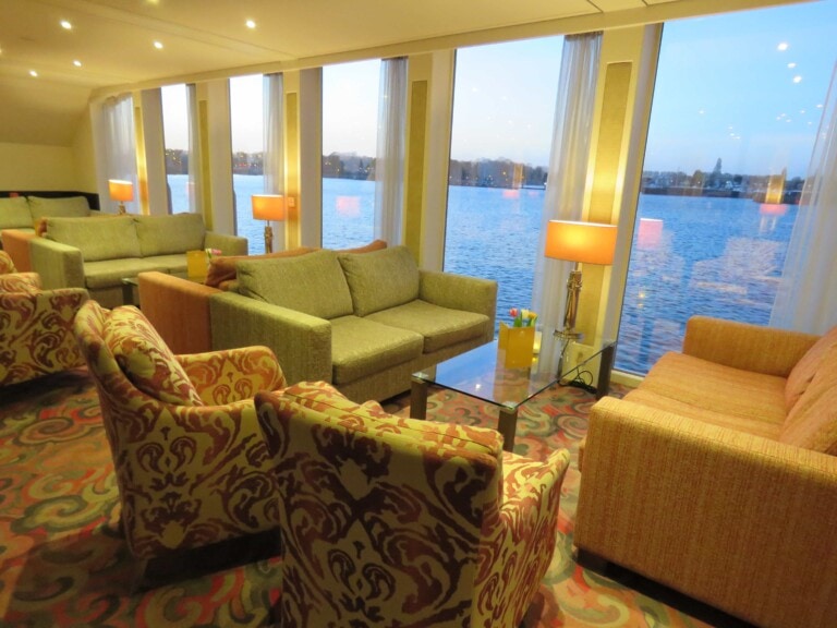 Modern lounge with armchairs and sofas arranged around glass tables, large windows overlooking a calm body of water at sunset, with lamps providing warm indoor lighting.