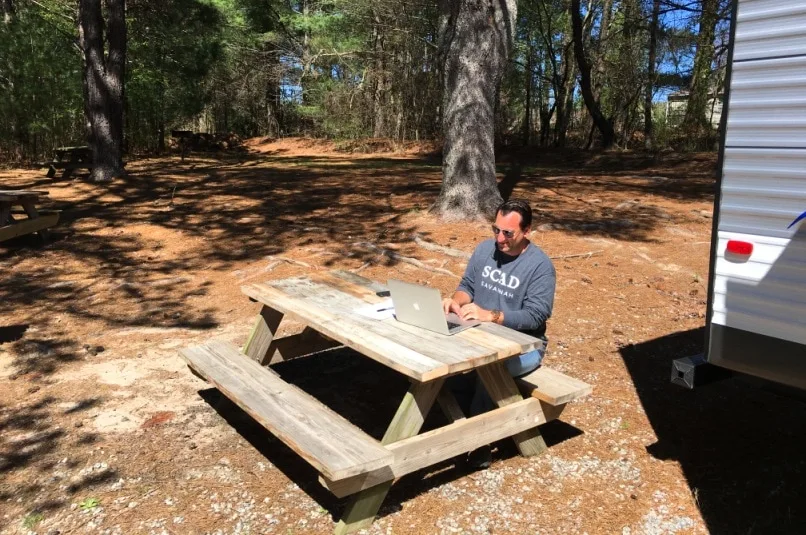 man sitting behind computer on wooden picnic table in the woods at RV park