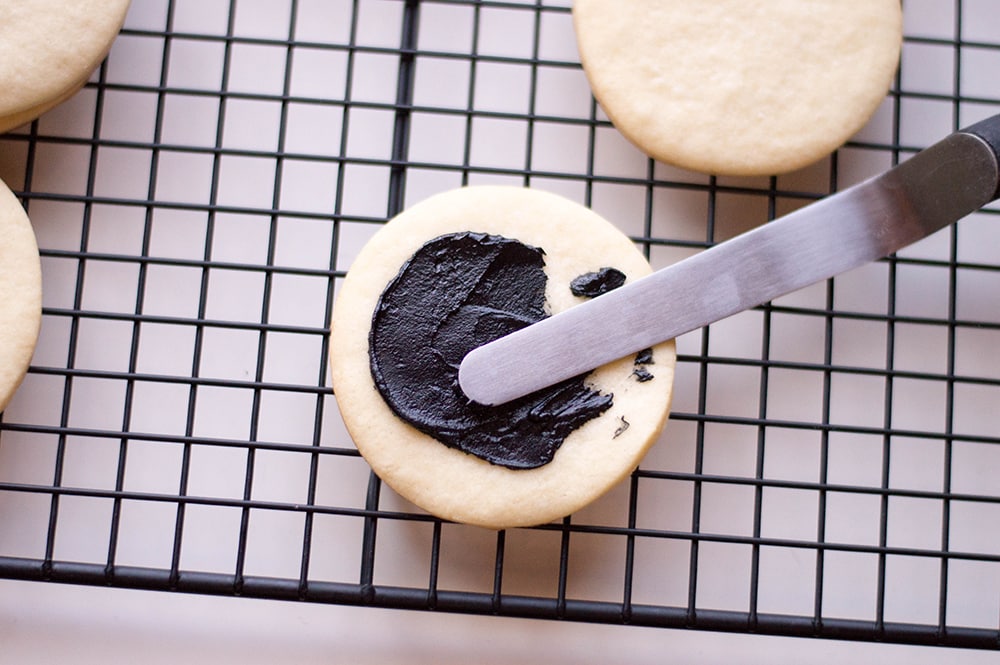 spreading black frosting on a sugar cookie on a wire rack