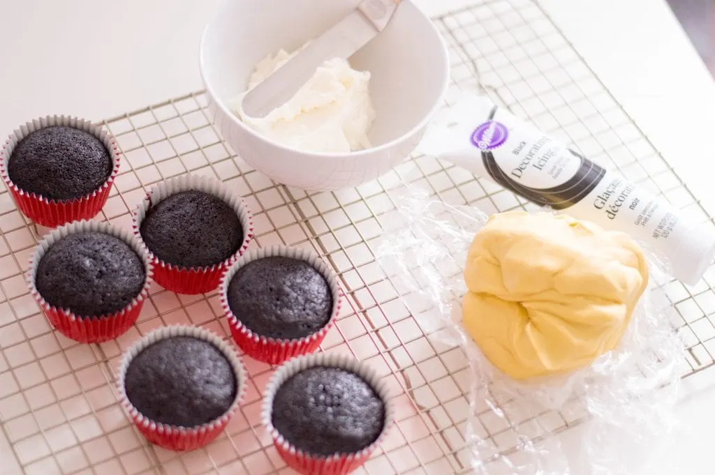 chocolate cupcakes in red wrappers with bowl of white frosting and yellow fondant