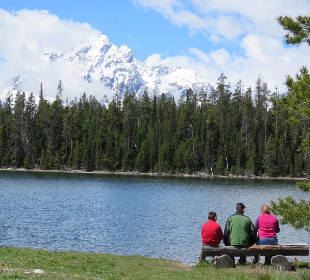 family sitting on a bench looking at Gran Teton Mountains across from lake