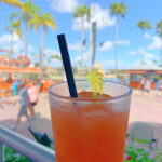 orange cocktail in a glass held in front of blue sky with palm trees in the distance
