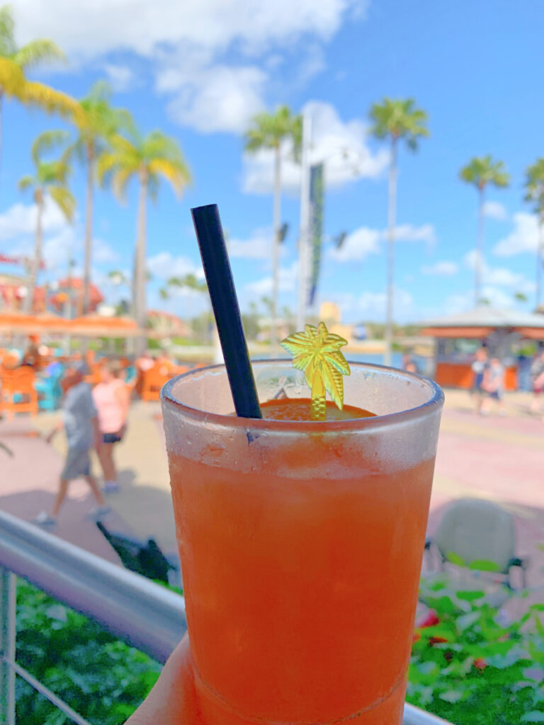 orange cocktail in a glass held in front of blue sky with palm trees in the distance