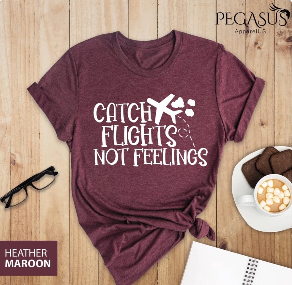 A heather maroon t-shirt with the text &ldquo;Catch Flights Not Feelings&rdquo; is laid on a wooden surface next to glasses, a notebook, chocolate cookies, and a mug of hot chocolate with marshmallows&mdash;making it a perfect choice for thoughtful flight attendant gifts.