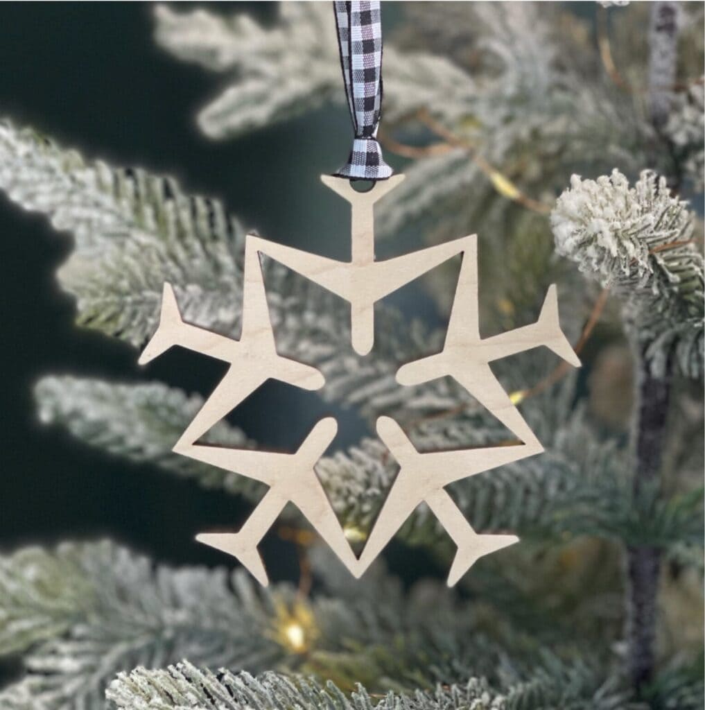 Wooden snowflake ornament with airplane-shaped arms hangs from a black-and-white checkered ribbon on a frosted Christmas tree branch&mdash;an ideal choice for unique flight attendant gifts.