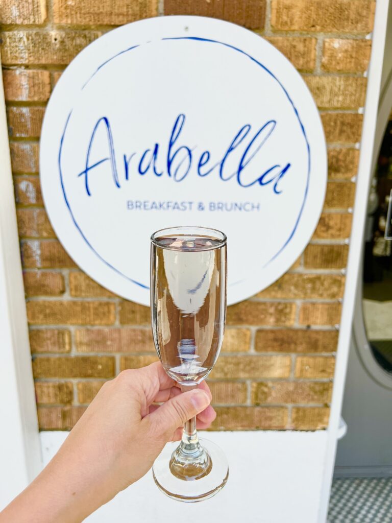 holding a glass of champagne in front of Arabella restaurant sign in Hendersonville