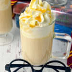 hot butterbeer drink in a glass mug topped with whipped cream and drizzled with butterscotch topping and black round glasses in foreground