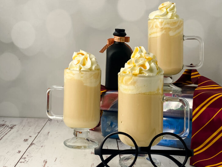 three glass mugs of hot butterbeer topped with whipped cream displayed on top of Harry Potter books with black round eyeglasses in foreground