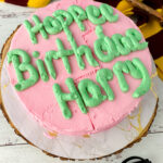 A pink and green decorated birthday cake with a Harry Potter scarf, wand, and glasses on the table for decoration.