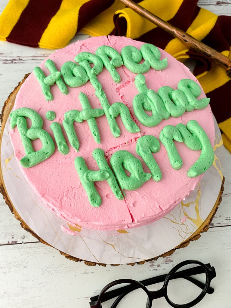 A pink and green decorated birthday cake with a Harry Potter scarf, wand, and glasses on the table for decoration.