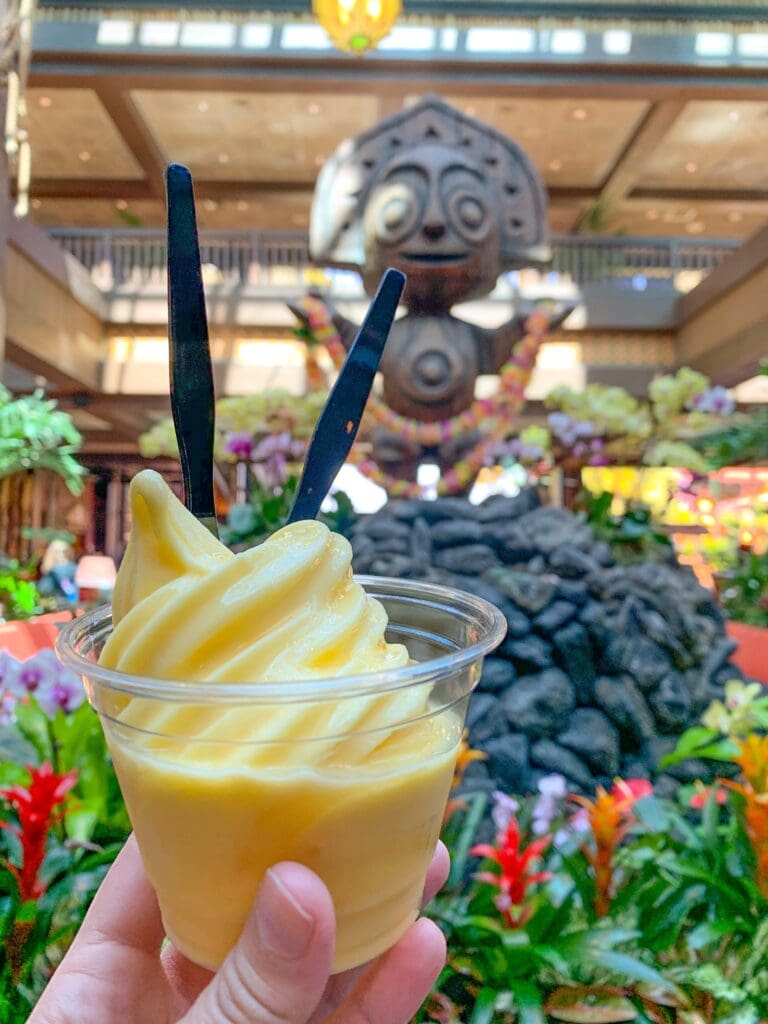 hand holding a dole whip in a plastic cup in front of Hawaiian statue
