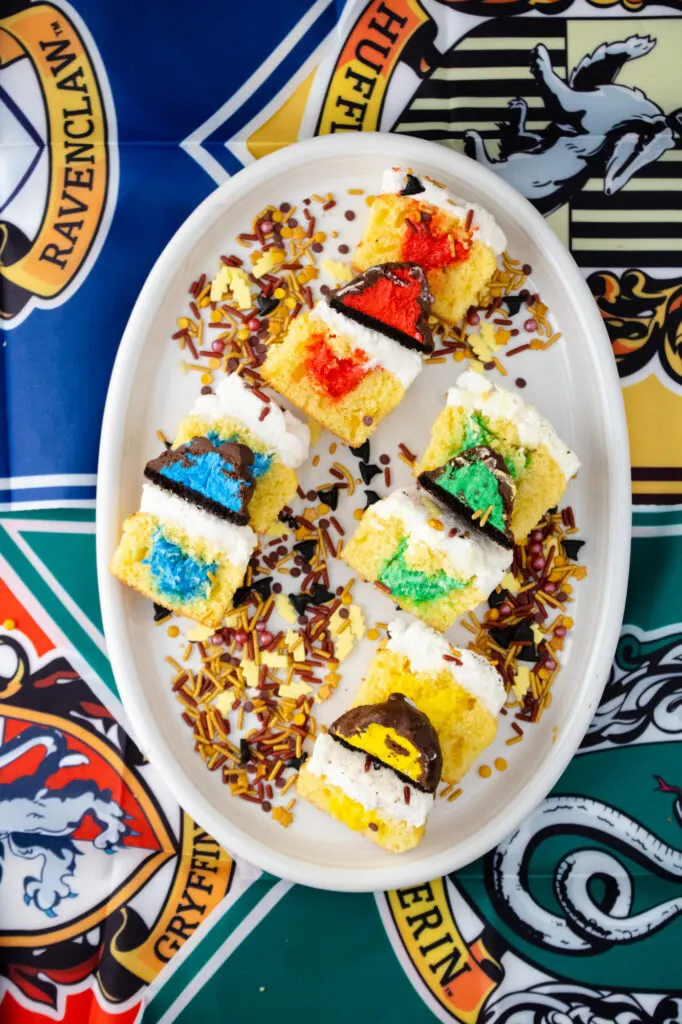 A white platter holds cupcakes sliced open to show blue, green, yellow, and red frostings; the sprinkled platter sits on a backdrop of Hogwarts house insignias.