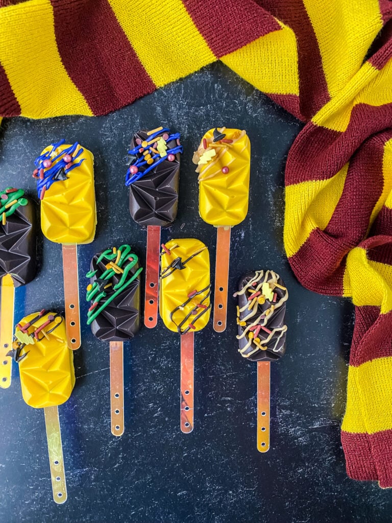 Drizzled and sprinkled yellow and black cakesicles laid out on a black speckled background with a red and yellow striped scarf.