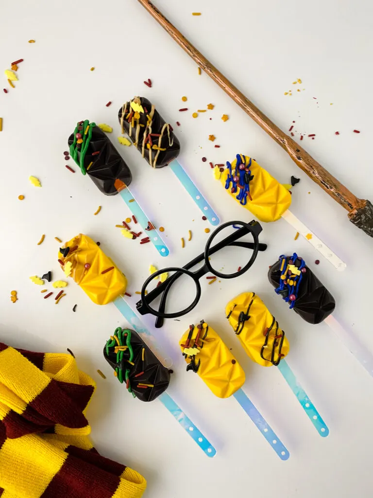 Drizzled and sprinkled yellow and black cakesicles laid out on a white background with a sorcerer's wand, Harry Potter glasses and striped scarf.