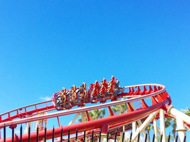 people in a roller coaster car moving along a curved red track with blue sky in background
