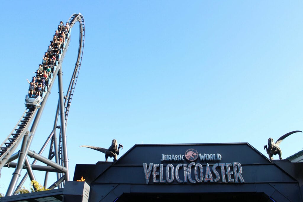 roller coaster ride high in the sky with people in a ride vehicle coming down the tracks and Jurassic Park VelociCoaster sign in foreground