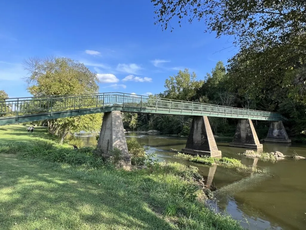 One of the many things to do in Asheboro is crossing a bridge over a river in a grassy area.